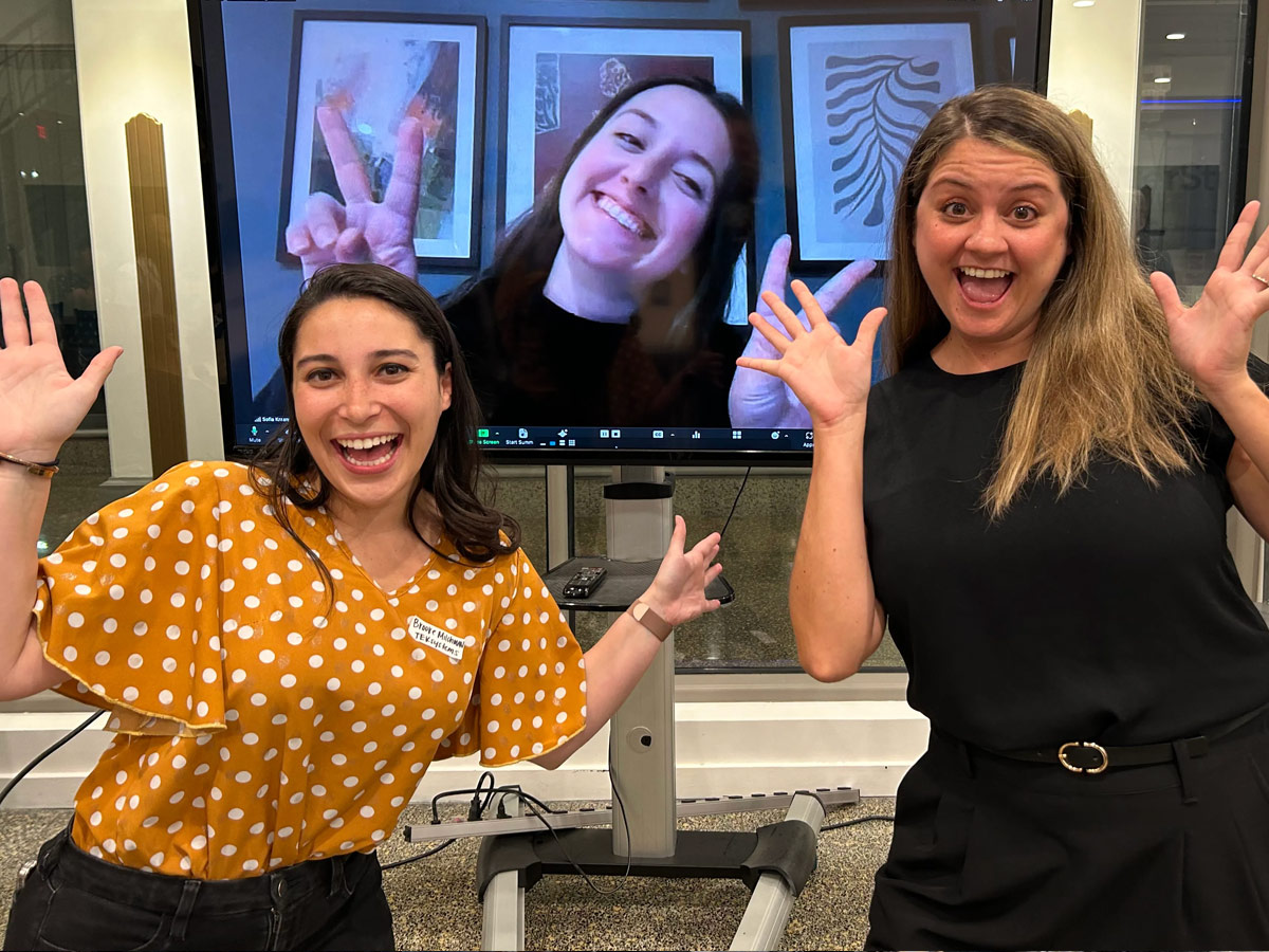 Three panelists from a recent meetup (two in-person, and one virtual on a TV in the background) pose for a photo with hands up.